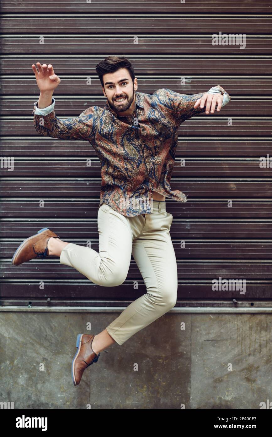 Junger bärtiger Mann im städtischen Hintergrund springen mit offenen Armen legere Kleidung. Kerl mit Bart und moderne Frisur in der Straße. Stockfoto
