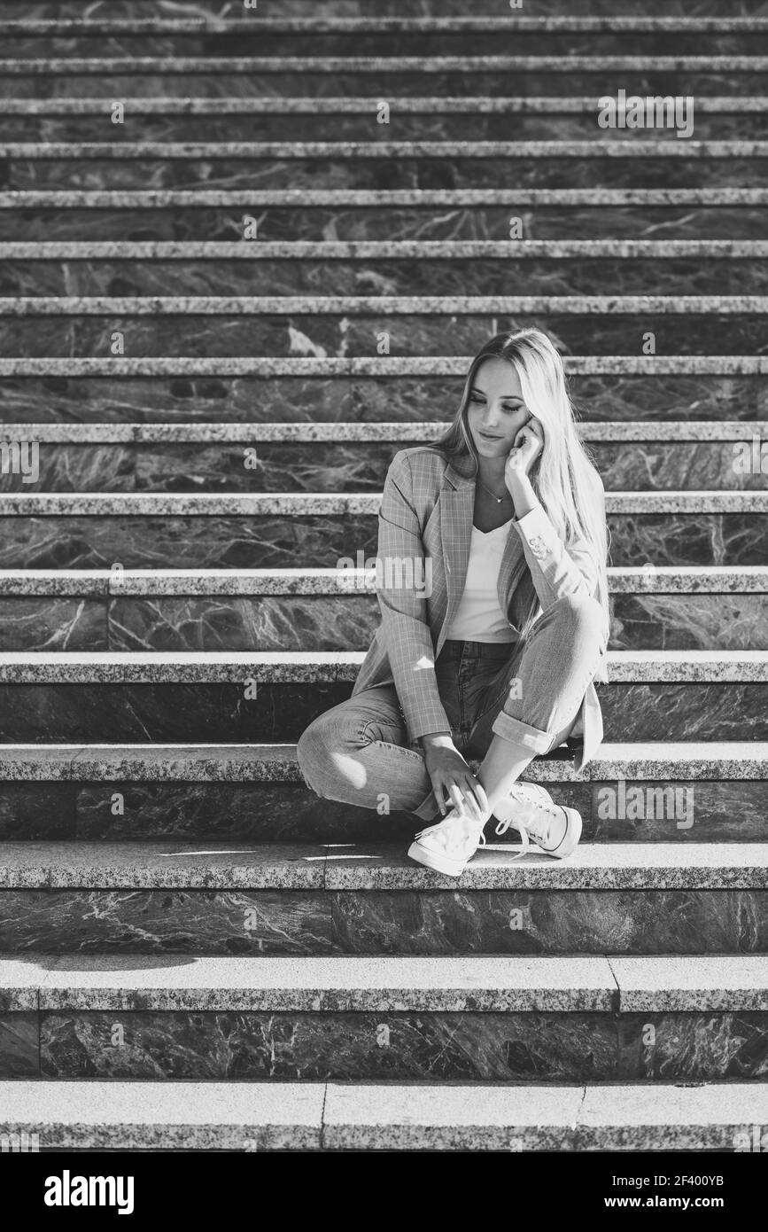 Nachdenklichen jungen kaukasischen Frau im städtischen Hintergrund. Blonde Mädchen legere Kleidung auf der Straße. Weibchen mit elegante Jacke und blaue Jeans sitzt auf der Treppe. Stockfoto