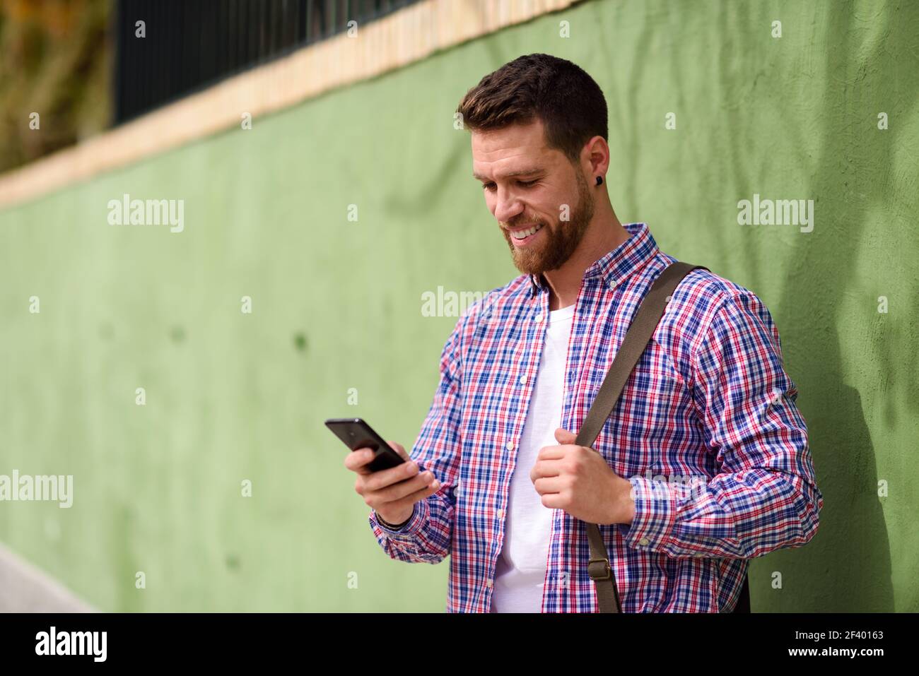Junger Mann, der sein Smartphone im urbanen Hintergrund betrachtet. Lebst. Junger Mann, der auf sein Smartphone schaut und im städtischen Hintergrund lächelt. Typ in legerer Kleidung. Lifestyle-Konzept. Stockfoto