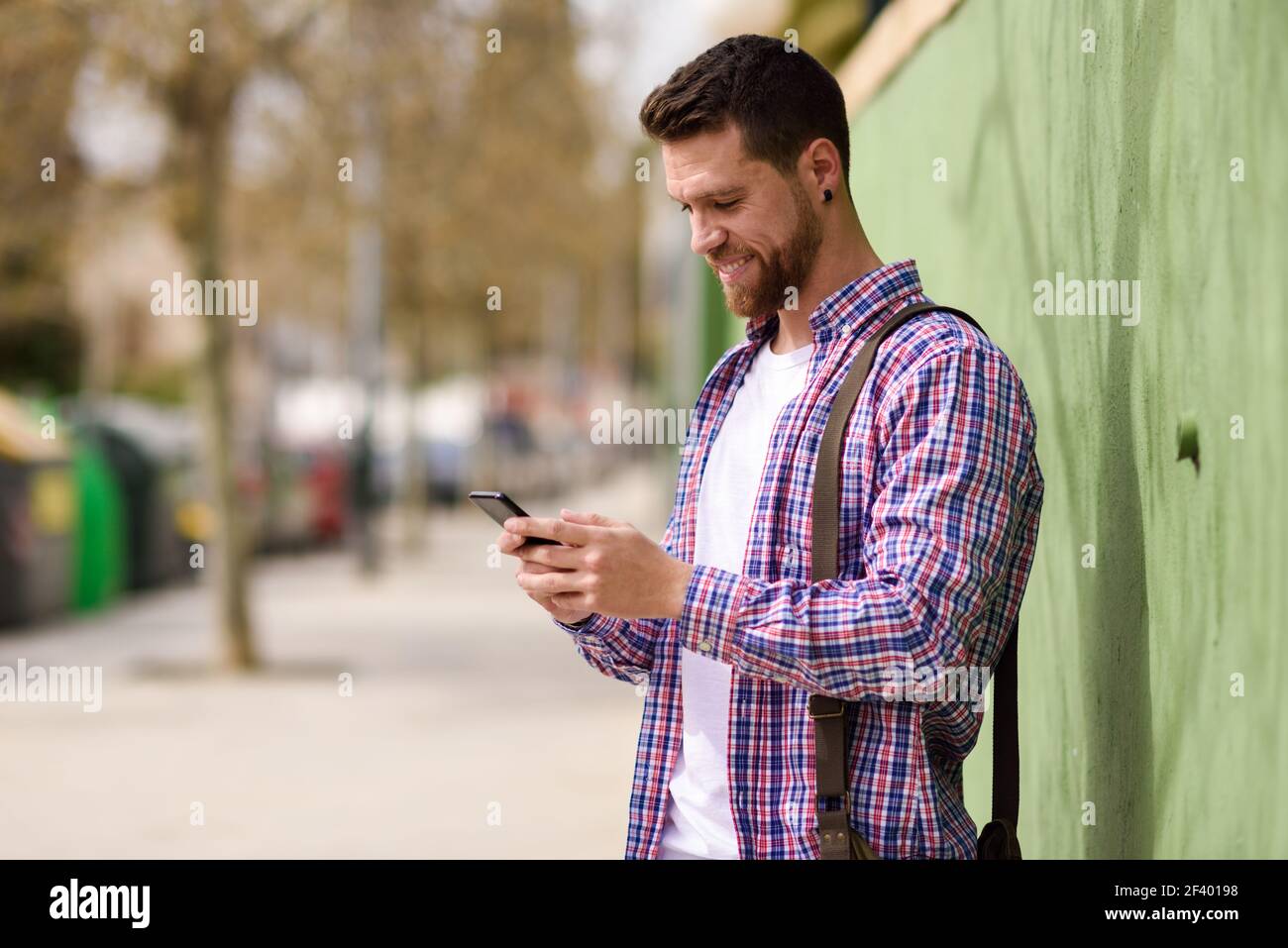 Lächelnder junger Mann, der sein Smartphone im städtischen Hintergrund sieht. L. lächelnder junger Mann, der sein Smartphone im städtischen Hintergrund sieht. Typ in legerer Kleidung. Lifestyle-Konzept. Stockfoto