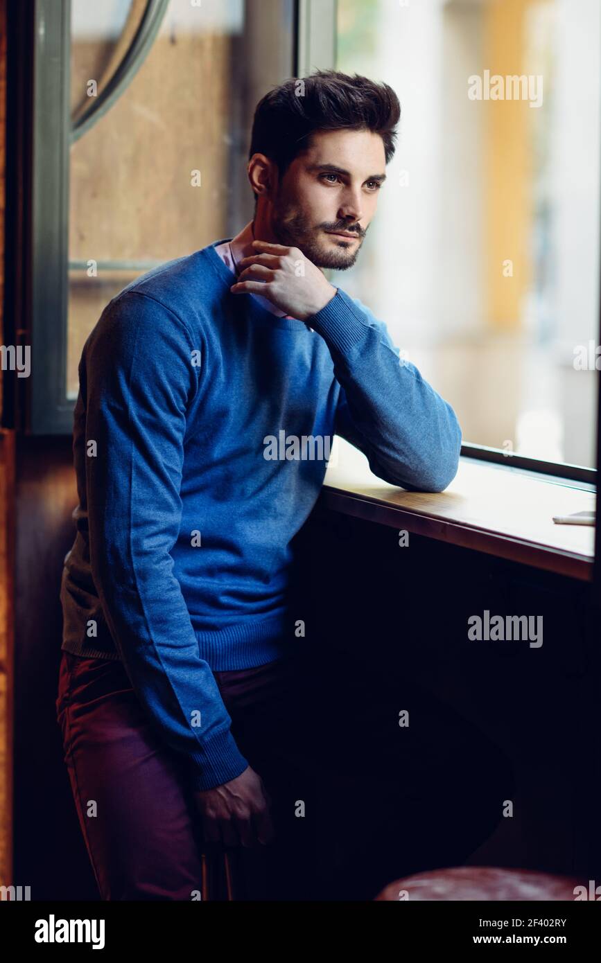 Nachdenklicher Mann mit blauen Pullover mit verlorenem Blick in der Nähe von einem Fenster in ein modernes Pub. Bärtiger Kerl mit modernen Frisur. Life style Konzept. Stockfoto
