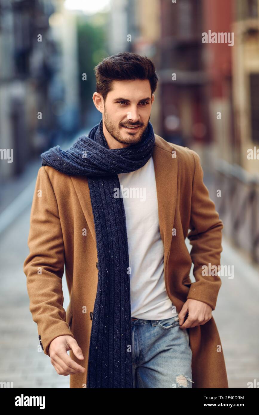 Junger Mann mit Winterkleidung in der Straße. Junge Bartgeier Kerl mit modernen Frisur mit Mantel, Schal, blaue Jeans und T-Shirt. Stockfoto