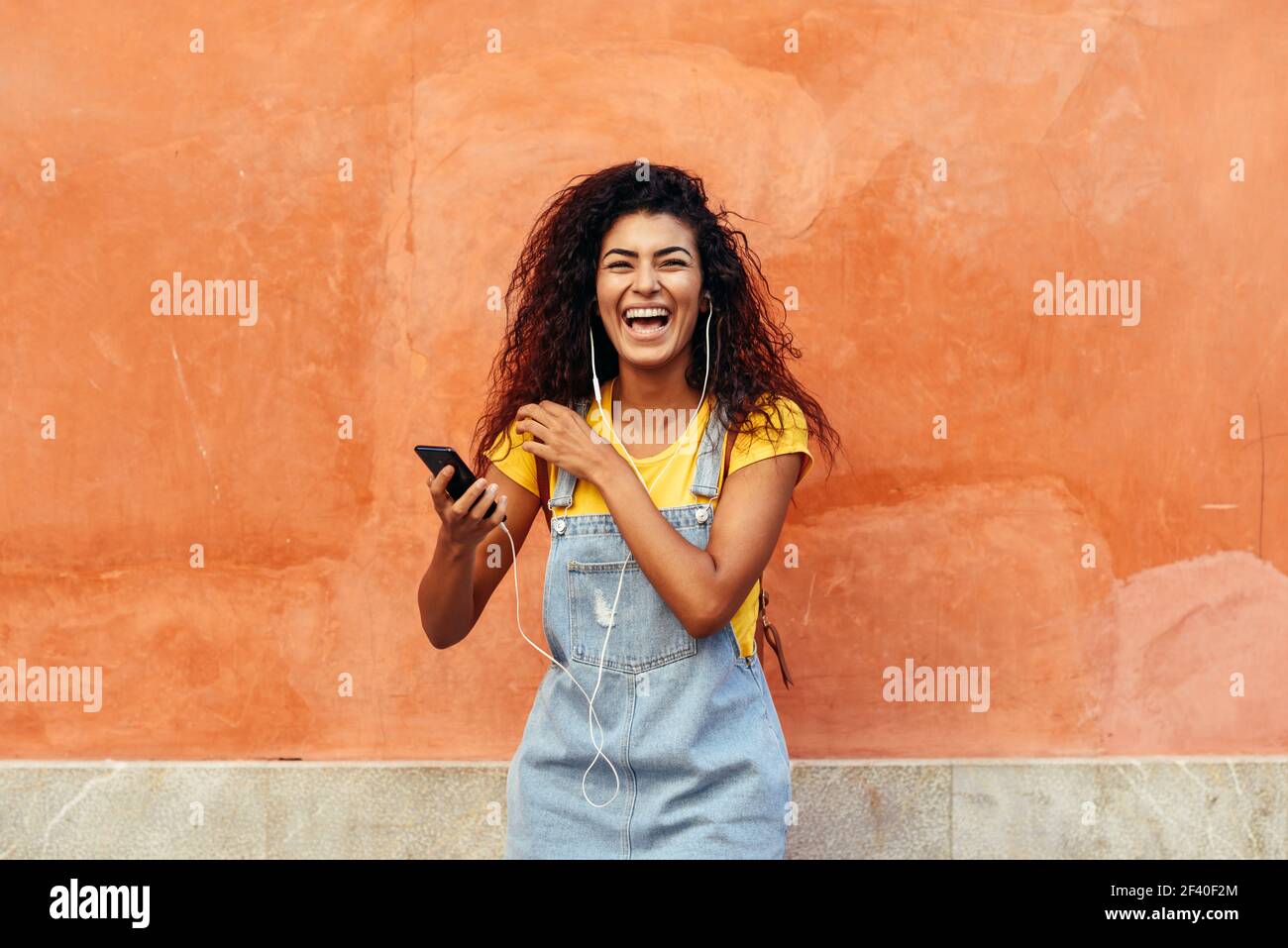 Junge schwarze Frau Lachen und Musik hören mit Kopfhörern im Freien. Arabische Mädchen in Freizeitkleidung mit Curly Frisur im städtischen Hintergrund. Stockfoto