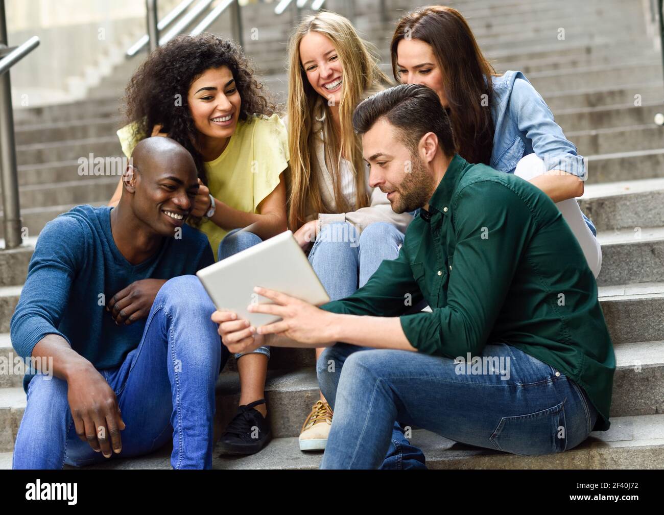 Multiethnische Gruppe von jungen Menschen, die einen Tablet-Computer im Freien im städtischen Hintergrund betrachten. Gruppe von Männern und Frauen sitzen zusammen auf Stufen.. Multiethnische Gruppe junger Menschen, die einen Tablet-Computer betrachten Stockfoto