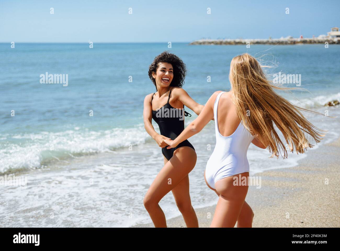 Zwei junge Frauen mit schönen Körpern in Bademode, die Spaß mit ihren  Händen am Strand gefangen haben. Lustige kaukasische und arabische Frauen  tragen schwarze und weiße Badeanzüge. Zwei Frauen im Badeanzug haben