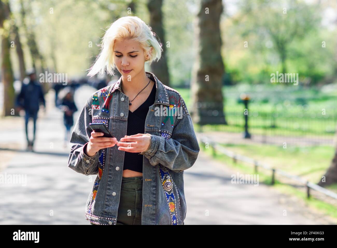 Junge urbane Frau mit moderner Frisur mit Smartphone zu Fuß in der Straße in einem Stadtpark in London, Großbritannien.. Junge urbane Frau mit moderner Frisur mit Smartphone zu Fuß in der Straße in einem Stadtpark. Stockfoto