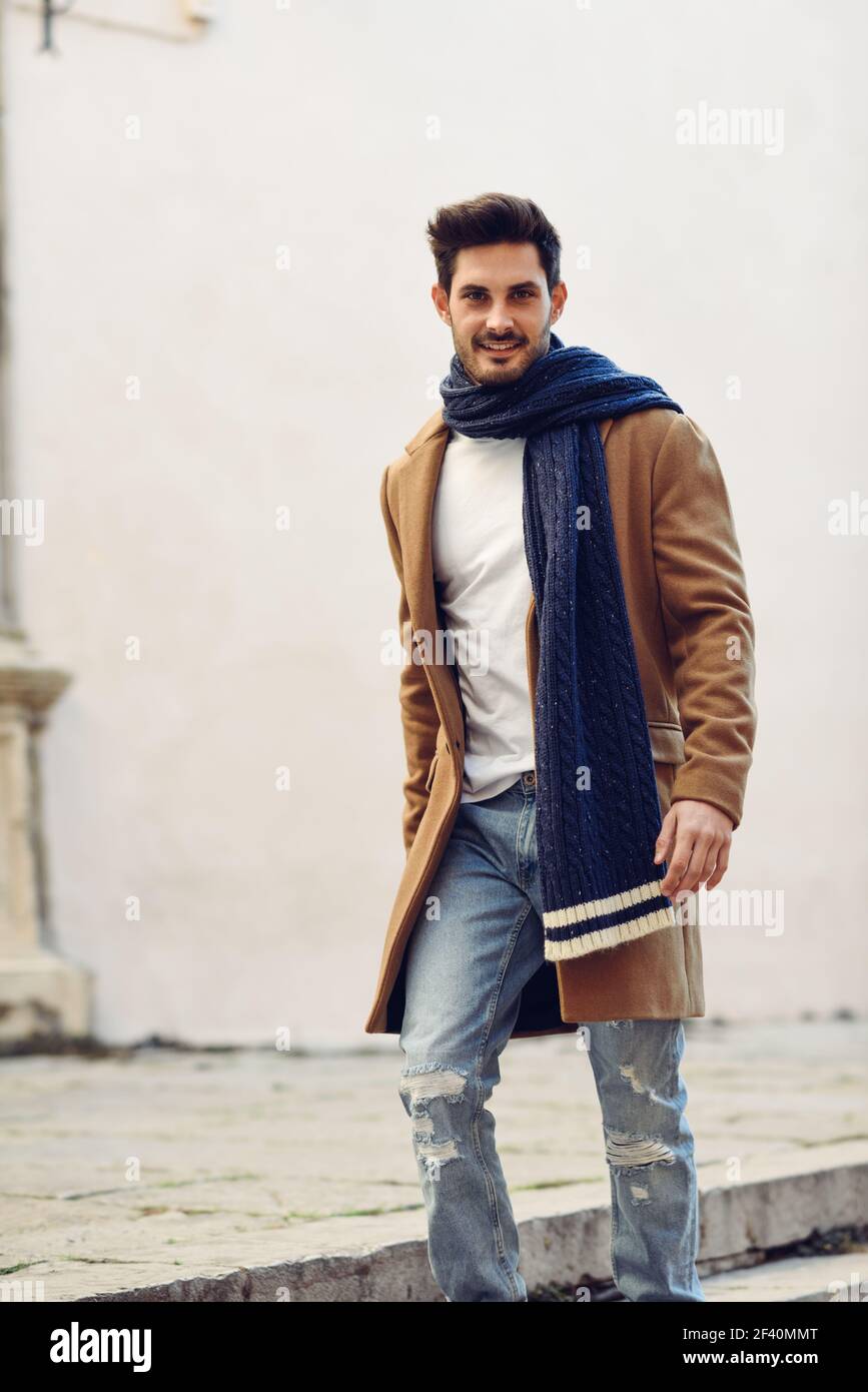 Junger Mann in Winterkleidung auf der Straße. Junger bärtiger Kerl mit moderner Frisur mit Mantel, Schal, blauen Jeans und T-Shirt. Junger Mann in Winterkleidung auf der Straße. Stockfoto