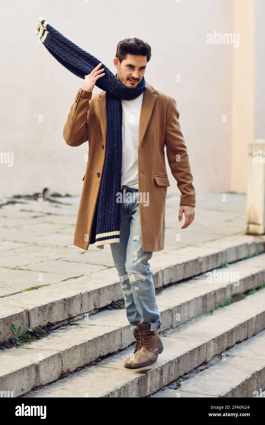Junger Mann in Winterkleidung auf der Straße. Junger bärtiger Kerl mit moderner Frisur mit Mantel, Schal, blauen Jeans und T-Shirt. Junger Mann in Winterkleidung auf der Straße. Stockfoto