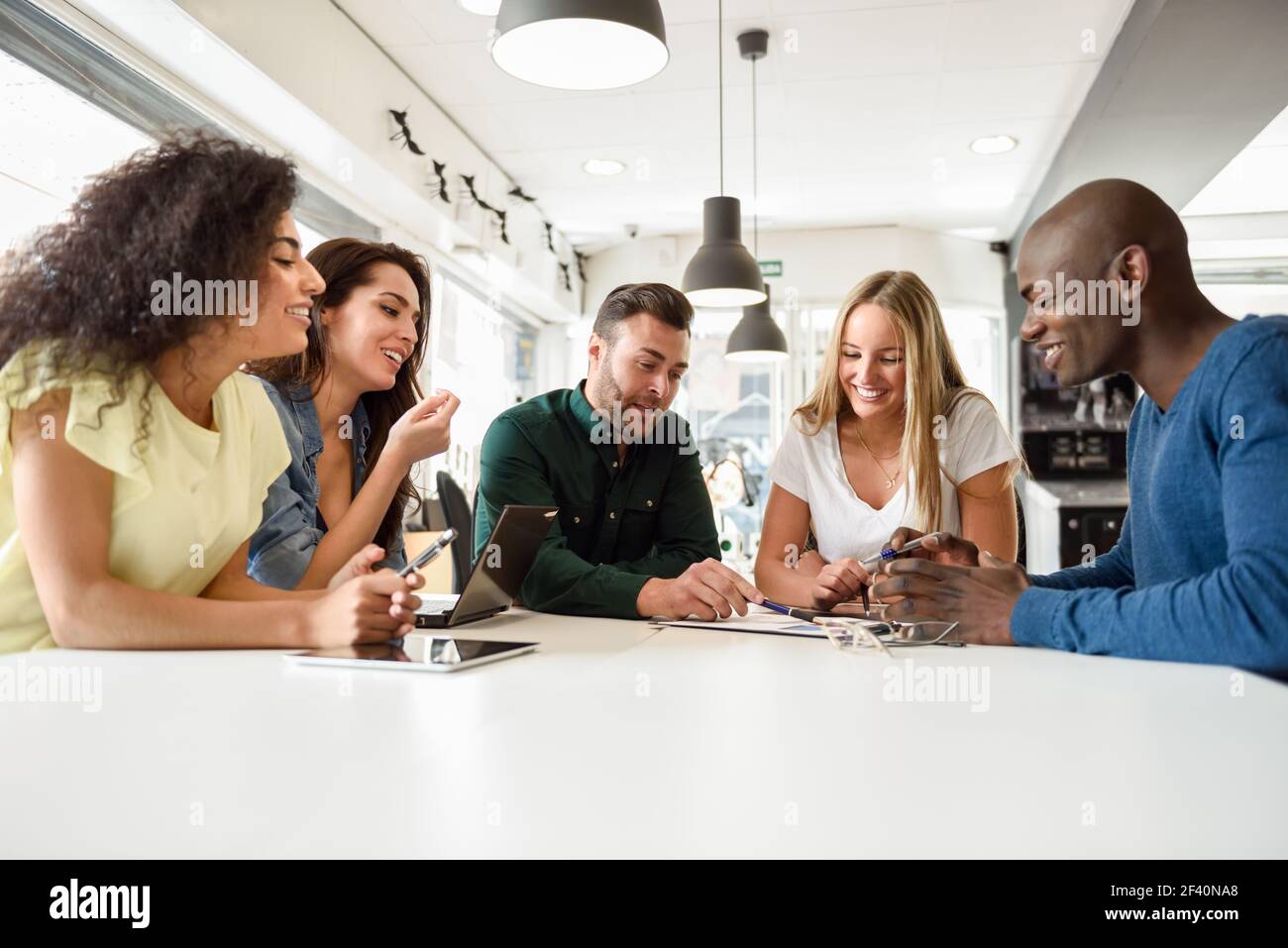 Fünf junge Menschen, die mit Laptop und Tablet auf weißem Schreibtisch studieren. Schöne Mädchen und Jungs arbeiten zusammen tragen legere Kleidung. Multi-ethnische Gruppe lächelt.. Multiethnische Gruppe von jungen Menschen, die gemeinsam am weißen Schreibtisch studieren Stockfoto
