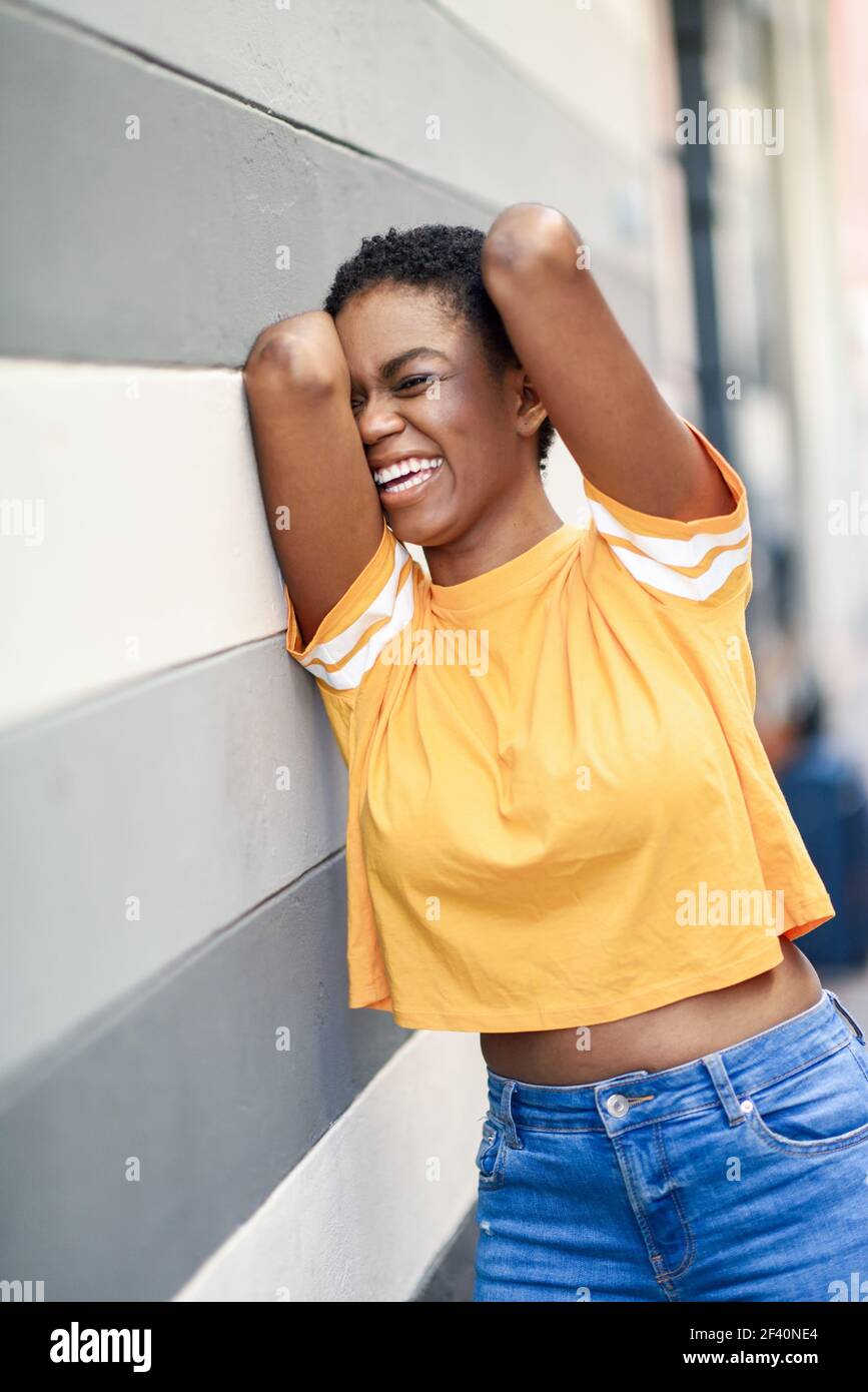 Junge Afrikanerin lachend nahe einer Stadtmauer. Mädchen mit sehr kurzen Haaren tragen lässige Kleidung.. Glückliche schwarze Frau lachend auf Stadtmauer. Stockfoto