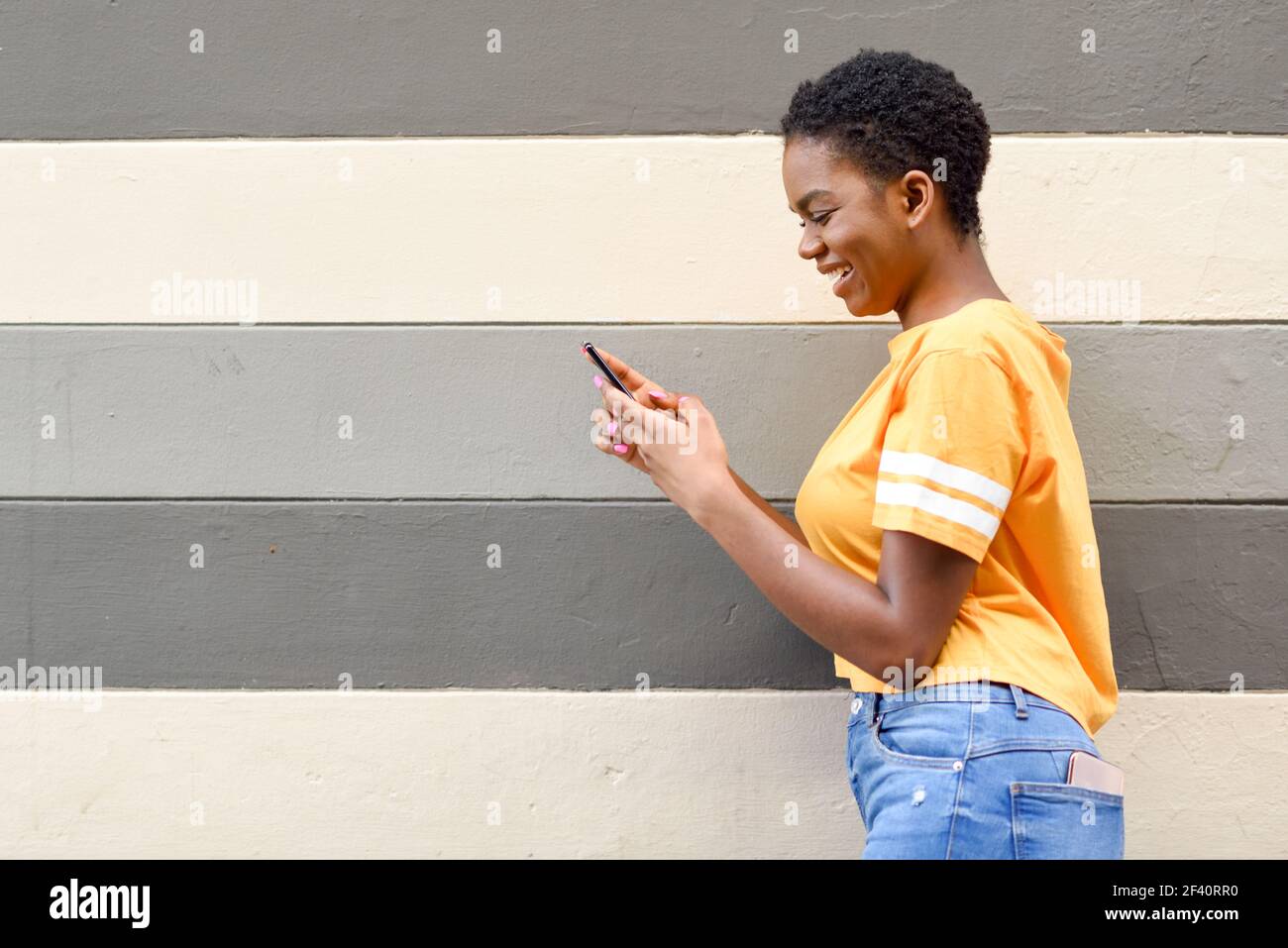 Junge schwarze Frau lacht mit ihrem Smartphone im Freien. Lifestyle-Konzept. Mädchen mit sehr kurzen Haaren tragen lässige Kleidung.. Junge schwarze Frau lacht mit ihrem Smartphone im Freien Stockfoto
