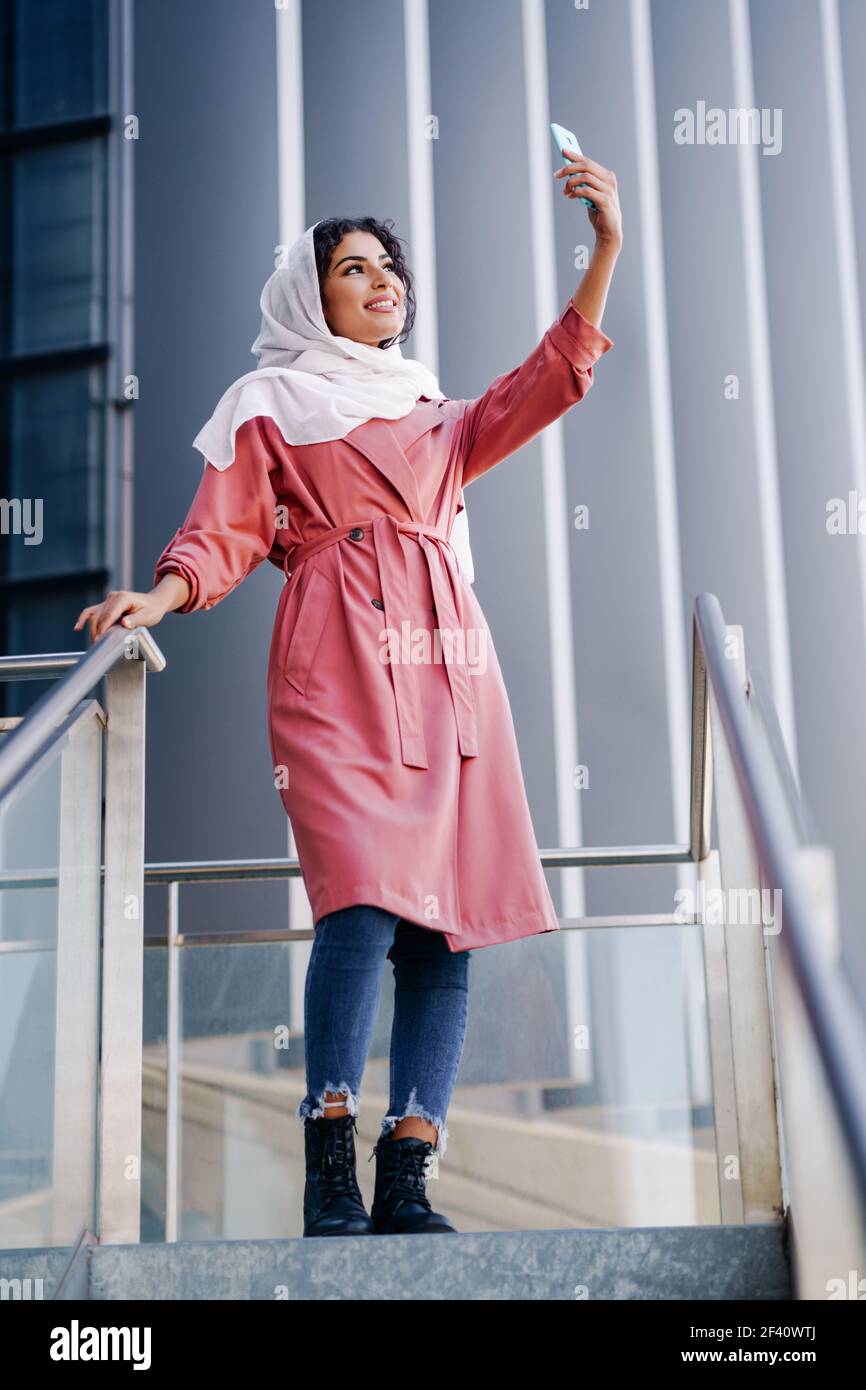 Junge arabische Frau trägt Hijab Kopftuch Foto mit einem Smartphone im städtischen Hintergrund.. Muslimische Frau mit Hijab nimmt Selfie mit Smartphone Stockfoto