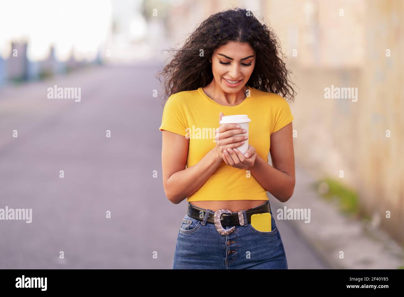 Junge arabische Frau, die mit einem Kaffee zum Mitnehmen über eine urbane Straße geht. Arabisches Mädchen, das mit einem Kaffee zum Mitnehmen über die Straße geht Stockfoto