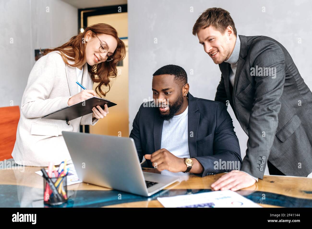 Lächelnde, multiethnische Kollegen arbeiten gemeinsam an einem neuen Projekt, mit einem Laptop. Erfolgreiche glückliche Geschäftspartner, die auf den Computerbildschirm schauen, im Internet surfen, im Büro sitzen Stockfoto
