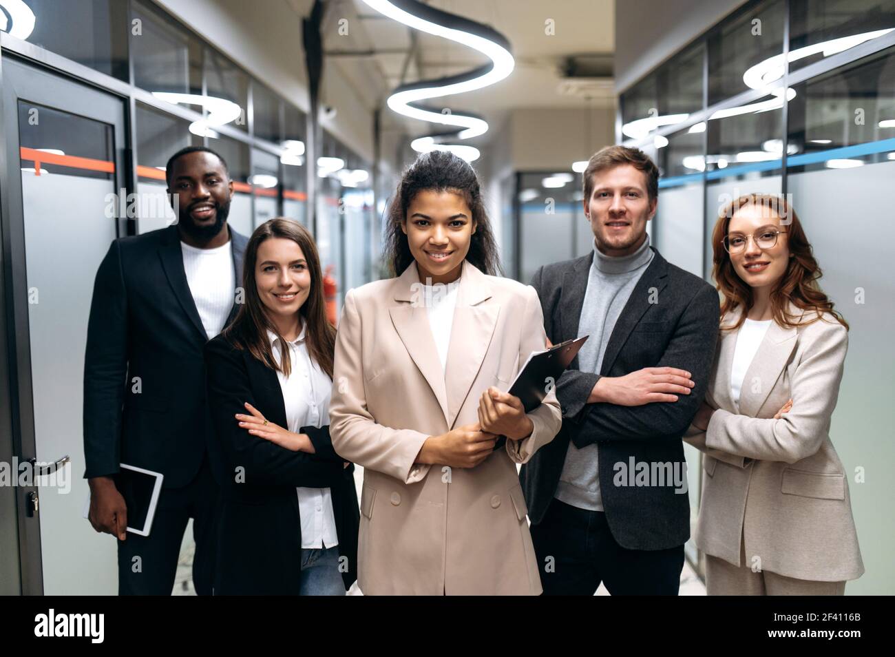 Erfolgreiche einflussreiche Unternehmer in stylischen Anzügen stehen im Büro mit Kreuzhänden. Glückliche multirassische Geschäftspartner, die selbstbewusst auf die Kamera blicken und lächeln Stockfoto