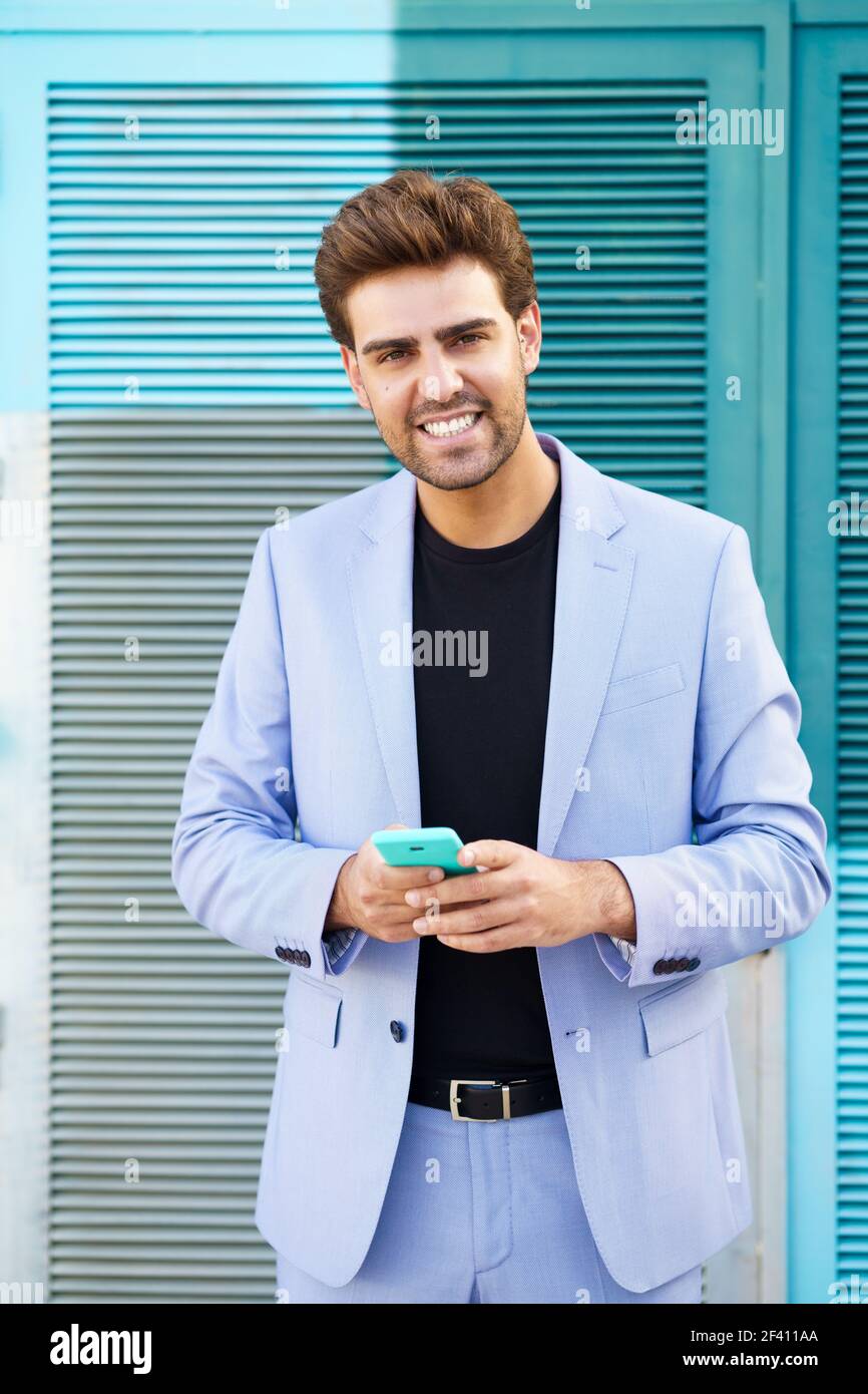 Junger Geschäftsmann trägt blauen Anzug mit einem Smartphone im städtischen Hintergrund. Mann mit formeller Kleidung auf der Straße. Junger Geschäftsmann trägt blauen Anzug mit einem Smartphone im städtischen Hintergrund. Stockfoto