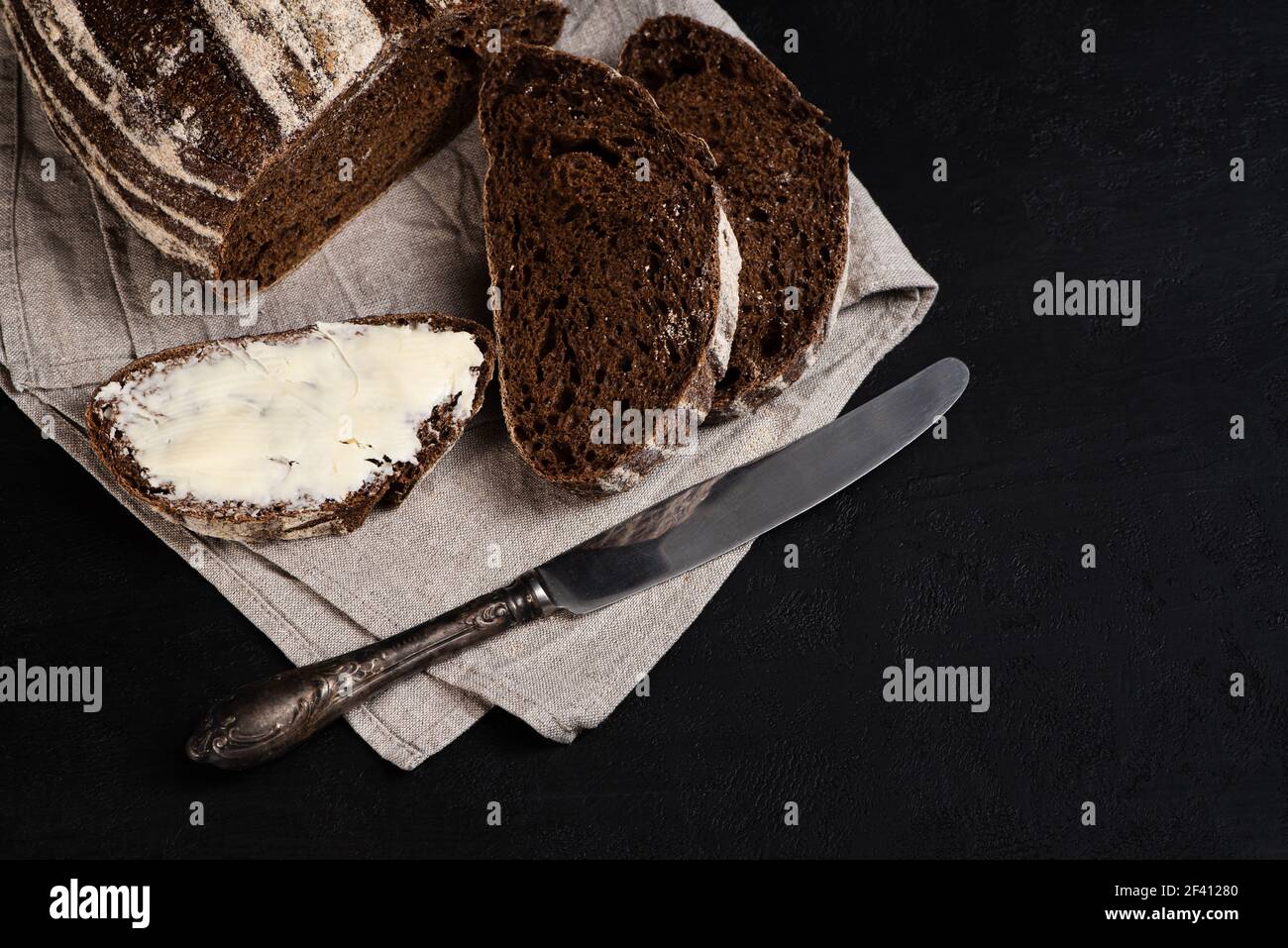 Gebutterte Scheibe frisch gebackenes Roggenbrot, Tischmesser auf dunklem Hintergrund. Handwerklich hergestelltes Sauerteig-Brot. Platz für Rezept kopieren. Blick von oben Stockfoto