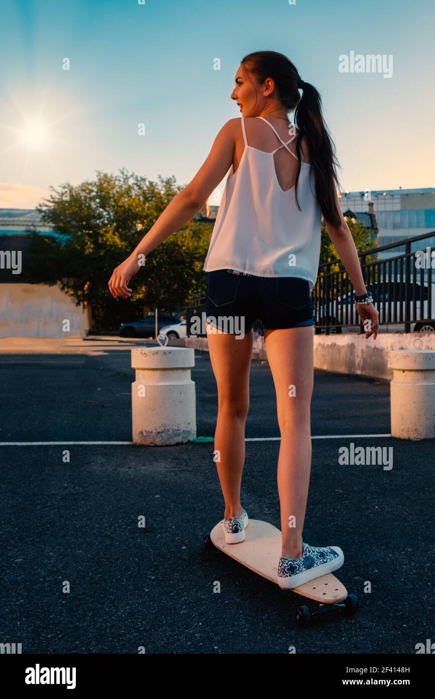 Schönes Mädchen Reiten Skateboard in kurzen Denim-Shorts, Blick von hinten vor dem frühen Abendhimmel. Schöne Mädchen Reiten Skateboard in kurzen Denim-Shorts, Blick von hinten Stockfoto