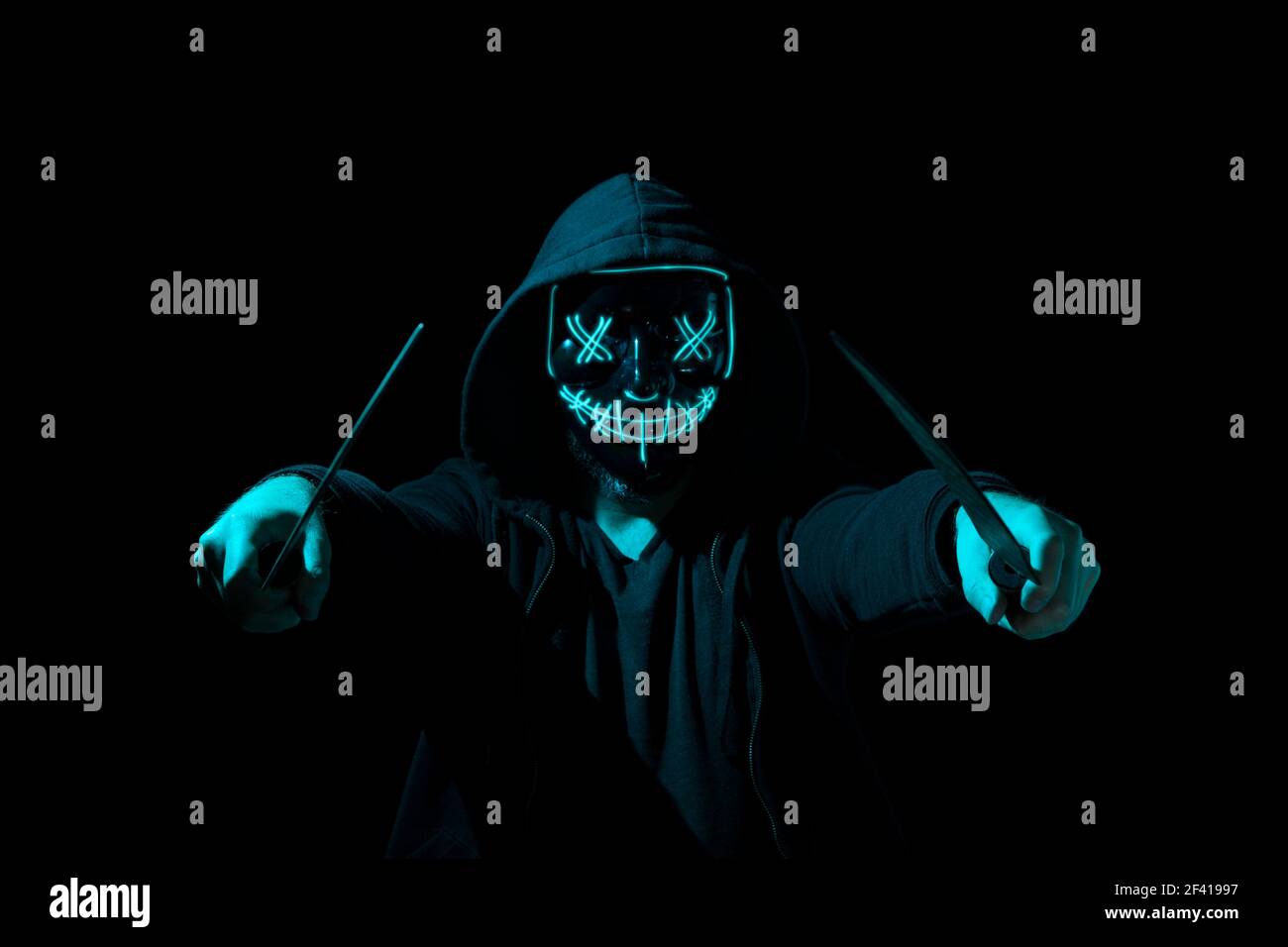 Mann bewaffnet mit zwei Macheten und trägt eine gruselige Beleuchtung Neon Glow Maske und ein Hoodie auf schwarzem Hintergrund. Stockfoto