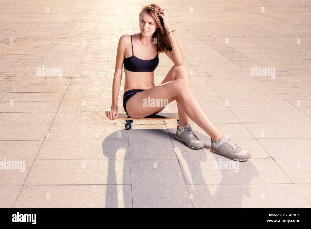 Schlanke sportliche Mädchen sitzt auf Skateboard in Fitness-Kleidung und Sneakers hinterleuchtet, viel Platz für Text. Slim sportliche Mädchen sitzt auf Skateboard in Fitness-Kleidung und Sneakers hinterleuchtet, Copyspace Stockfoto