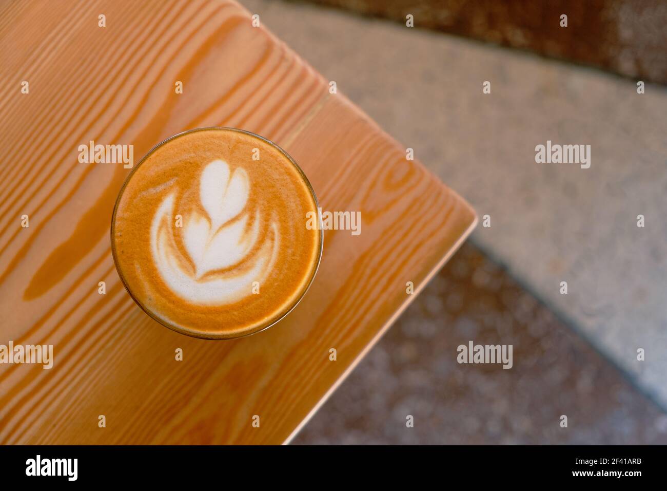 Flacher weißer Kaffee auf der Tischecke von oben. Flacher weißer Kaffee in transparentem Glas in einem Café.Es ist Espresso-basierte Kaffeegetränk bestehend aus Espresso mit Mikroschaum (gedämpfte Milch mit feinen Blasen). Flacher weißer Kaffee auf der Tischecke von oben. Flacher weißer Kaffee aus transparentem Glas. Es handelt sich um ein Kaffeegetränk auf Espresso-Basis, bestehend aus Espresso mit Mikroschaum (gedämpfte Milch mit kleinen, feinen Blasen). Stockfoto