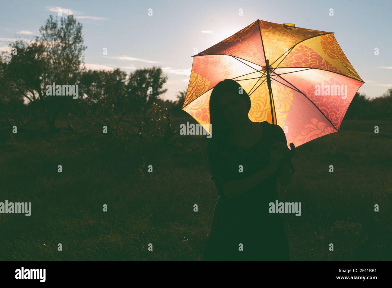 Ein Mädchen mit einem farbigen Regenschirm posiert auf einem Feld. Der Schirm wird von hinten durch das Sonnenuntergangslicht beleuchtet. Es&rsquo;s Raum auf der linken Seite für einen Copyspace.. Ein Mädchen mit einem farbigen Regenschirm posiert auf einem Feld. Der Schirm wird von hinten durch das Sonnenuntergangslicht beleuchtet Stockfoto