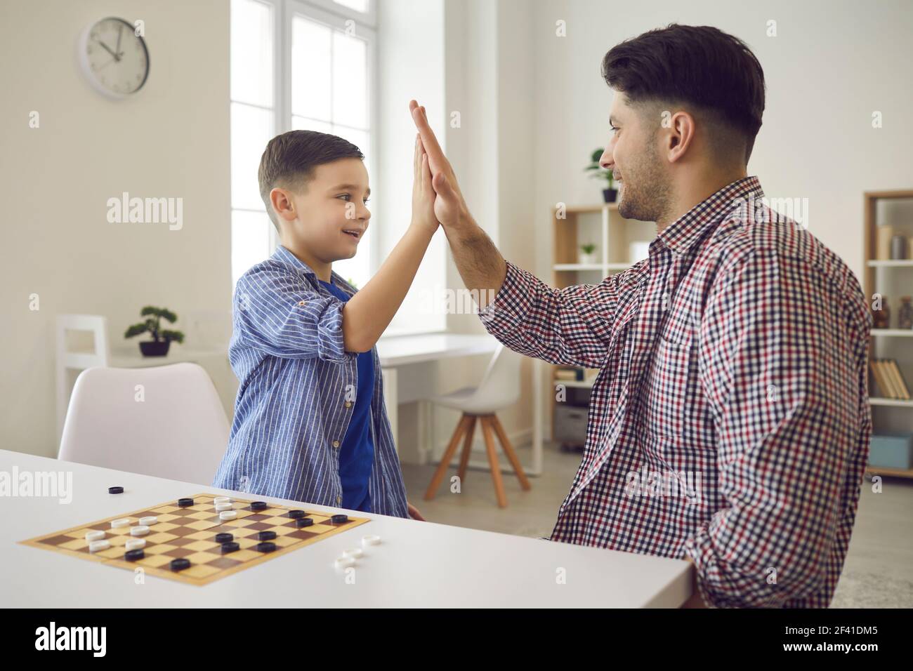 Fröhlicher kleiner Junge gibt seinem Vater fünf, während er zu Hause Schach spielt. Papa verbringt Zeit mit seinem Sohn. Stockfoto