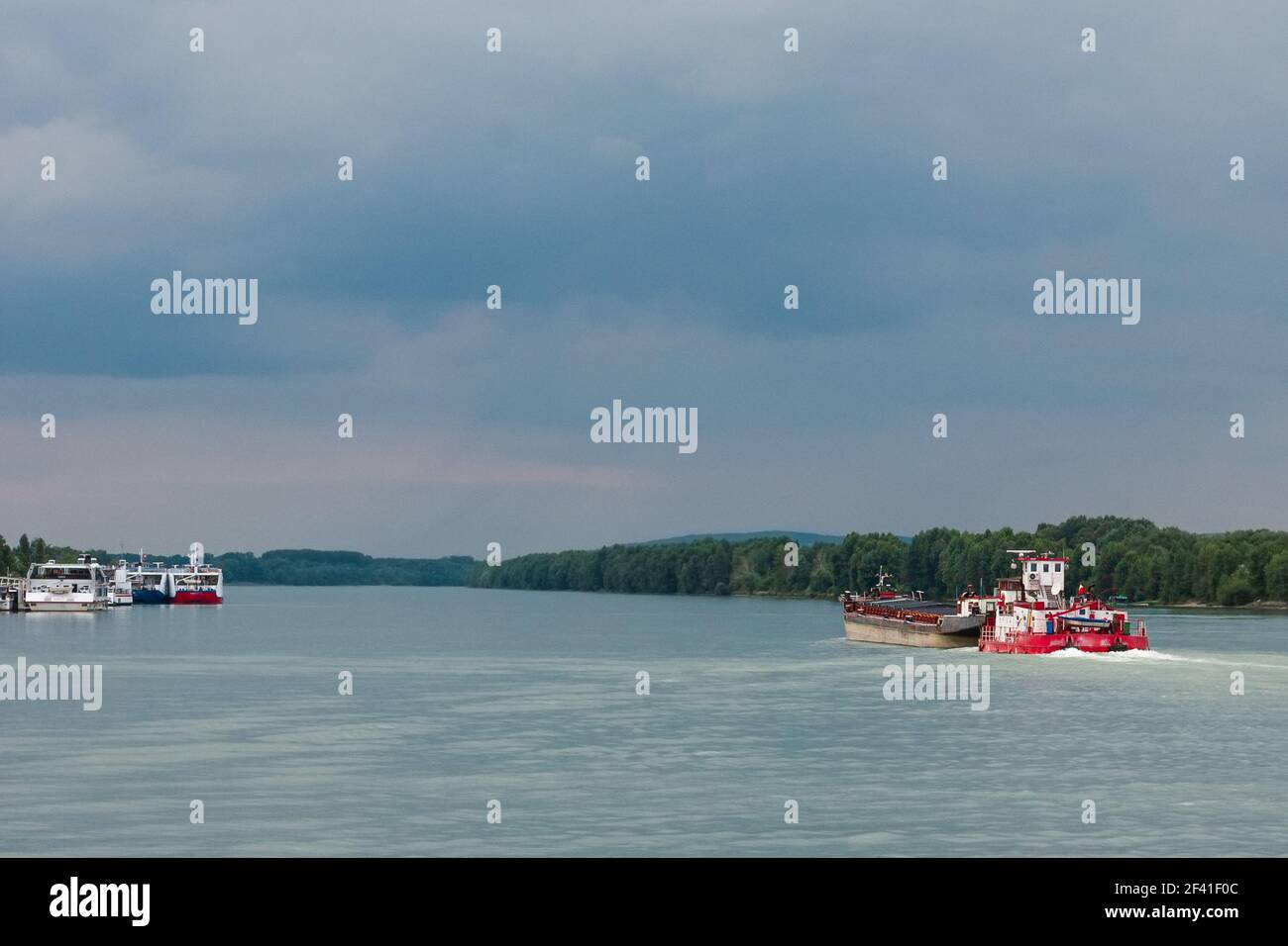 Rote, weiße Frachtschiffe segeln bei bewölktem Wetter entlang des breiten Flussufers, geparkte Schiffe sind in der Ferne. Selektiver Fokus. Barge auf der rechten Seite ist Segel Stockfoto