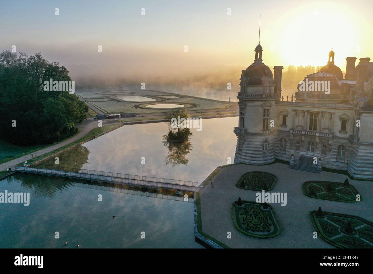 Morgenblick auf die Wasseroberfläche und die Schlossgärten im Nebel. Erhabener Blick gegen aufgehende Sonne, Silhouette des Schlosses Chantilly, Oise, Frankreich. Stockfoto