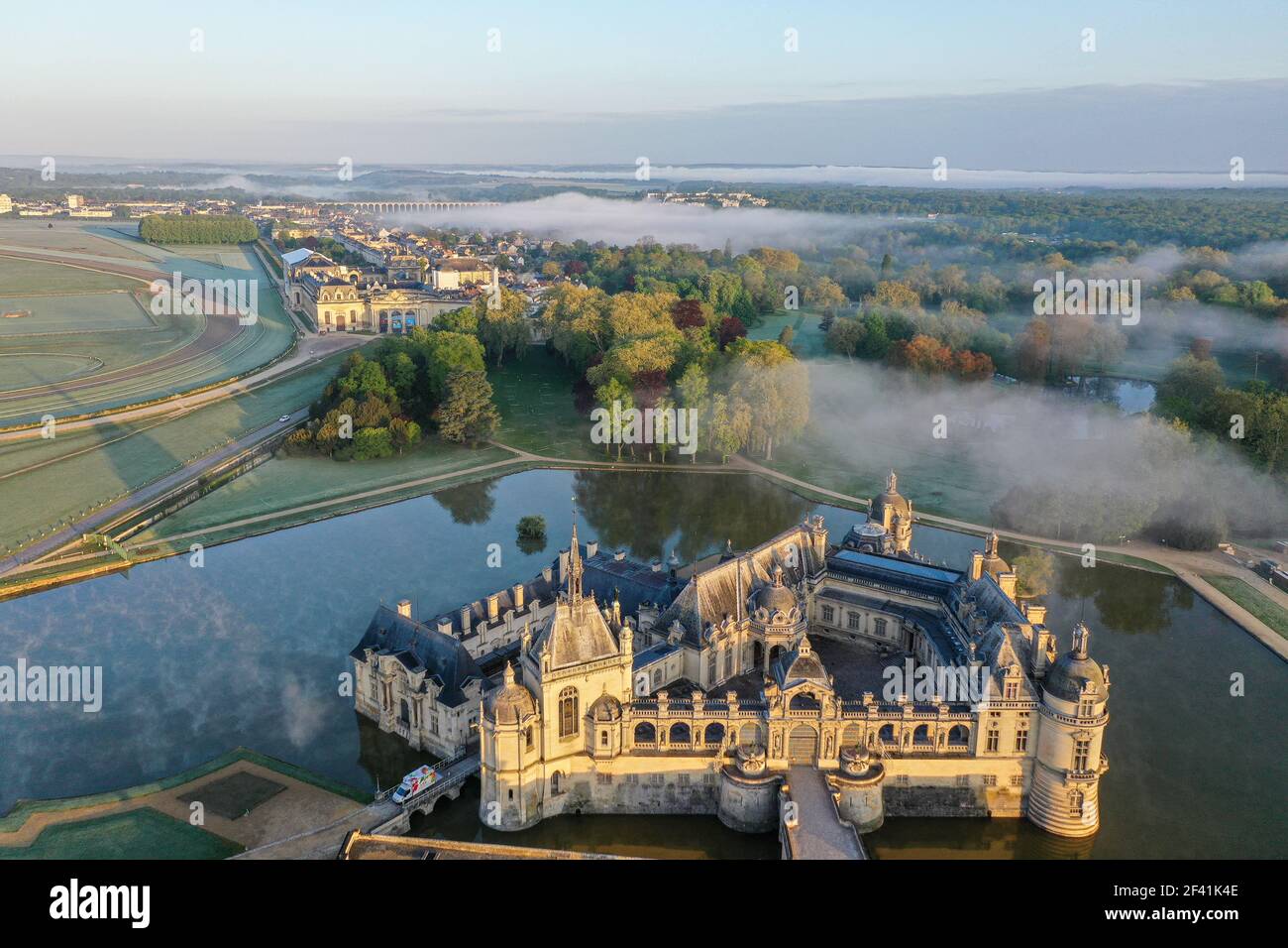 Drohnenansicht des historischen Schlosses Chateau de Chantilly, das morgens von hellem Sonnenlicht beleuchtet wird. Spärlicher Nebel bleibt über Wasseroberflächen. Stockfoto