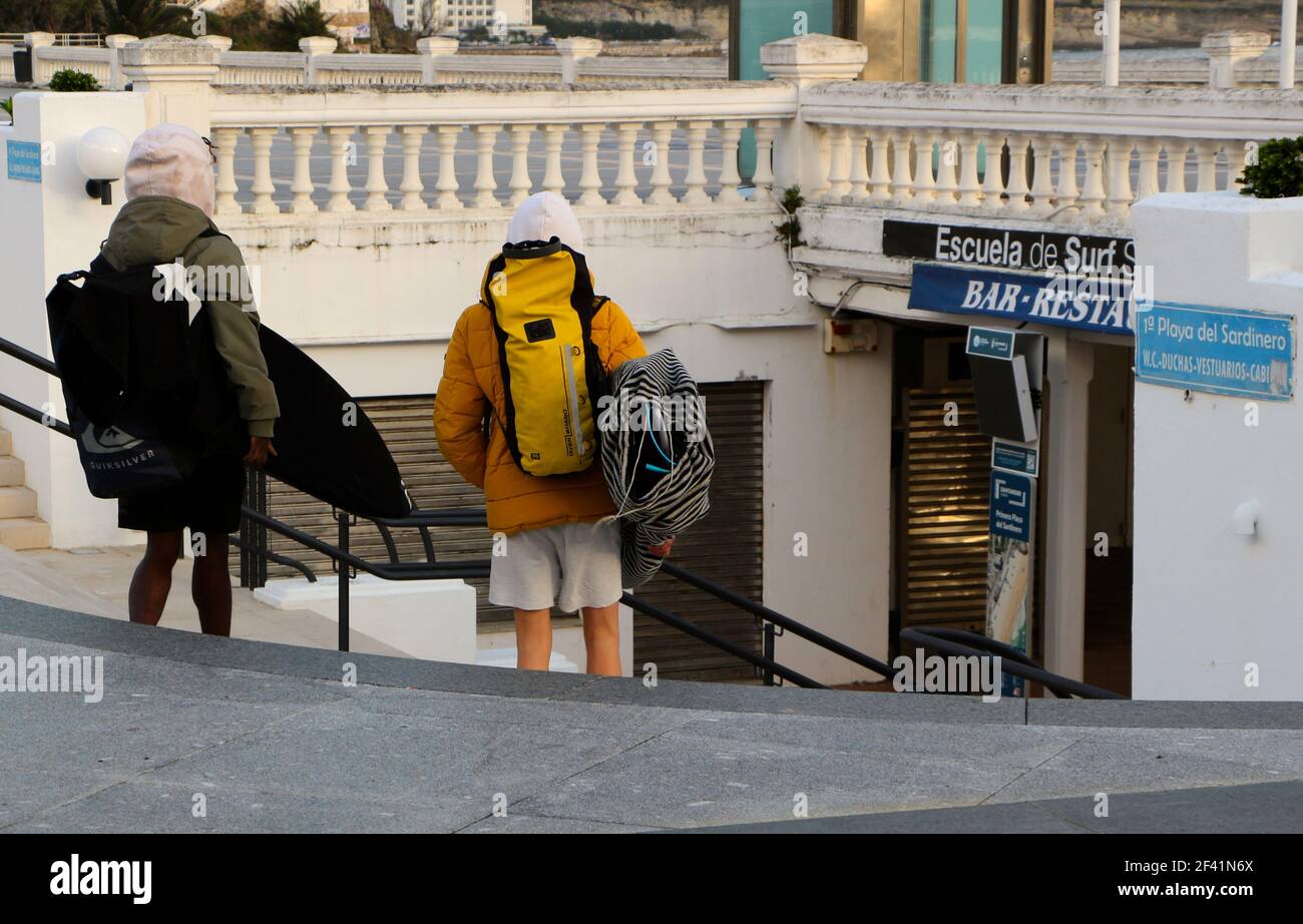 Zwei junge Männer, die Surfbretter an einem windigen Wintermorgen auf dem Weg zu einer Surfschule in Sardinero Santander Cantabria Spanien tragen Stockfoto