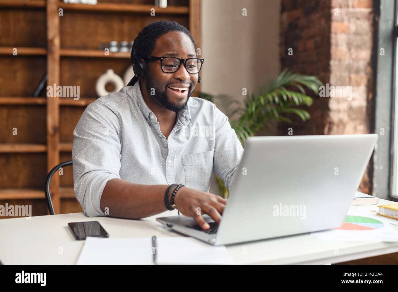 Fokussierter afroamerikanischer Geschäftsmann in Brillen, der Kopfhörer trägt, Videoanruf mit Kunden auf Laptop hält, konzentrierter Mann in Brillen, der Online-Unterricht hält, Kunde berät Stockfoto