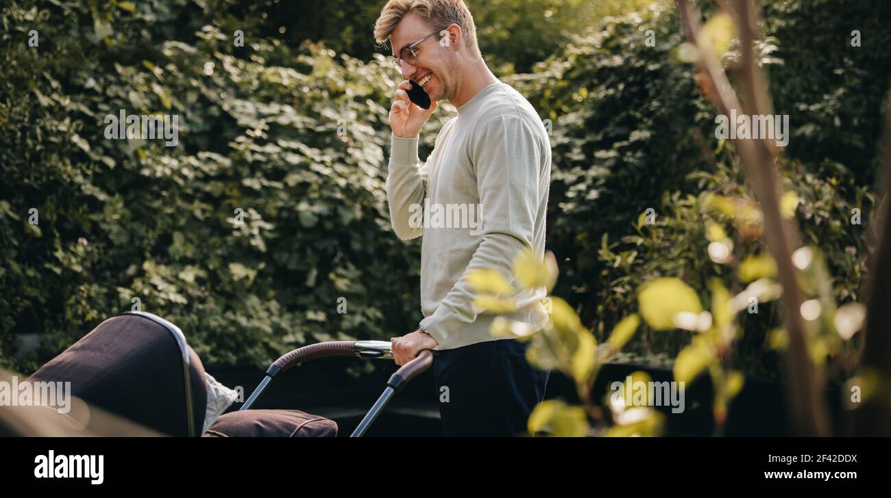 Mann, der mit dem Handy spricht und den Kinderwagen nach draußen schiebt. Vater auf Vaterschaftsurlaub. Stockfoto