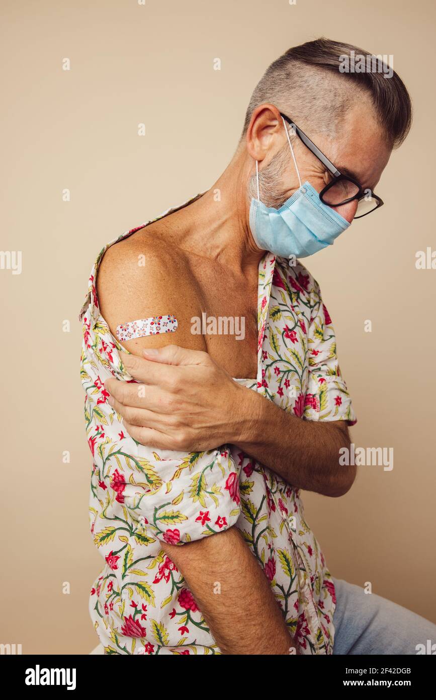 Reifer Mann trägt eine schützende Gesichtsmaske mit Verband am Arm nach dem Impfen. Mittelalter Mann nach Erhalt der Immunitätsimpfung mit Blick nach unten. Stockfoto