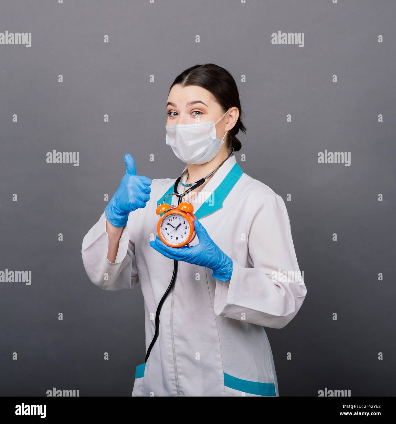 Lächelnde Ärztin Frau in Maske zeigt auf Uhr, Zeit zu impfen, medizinische Konzept Stockfoto