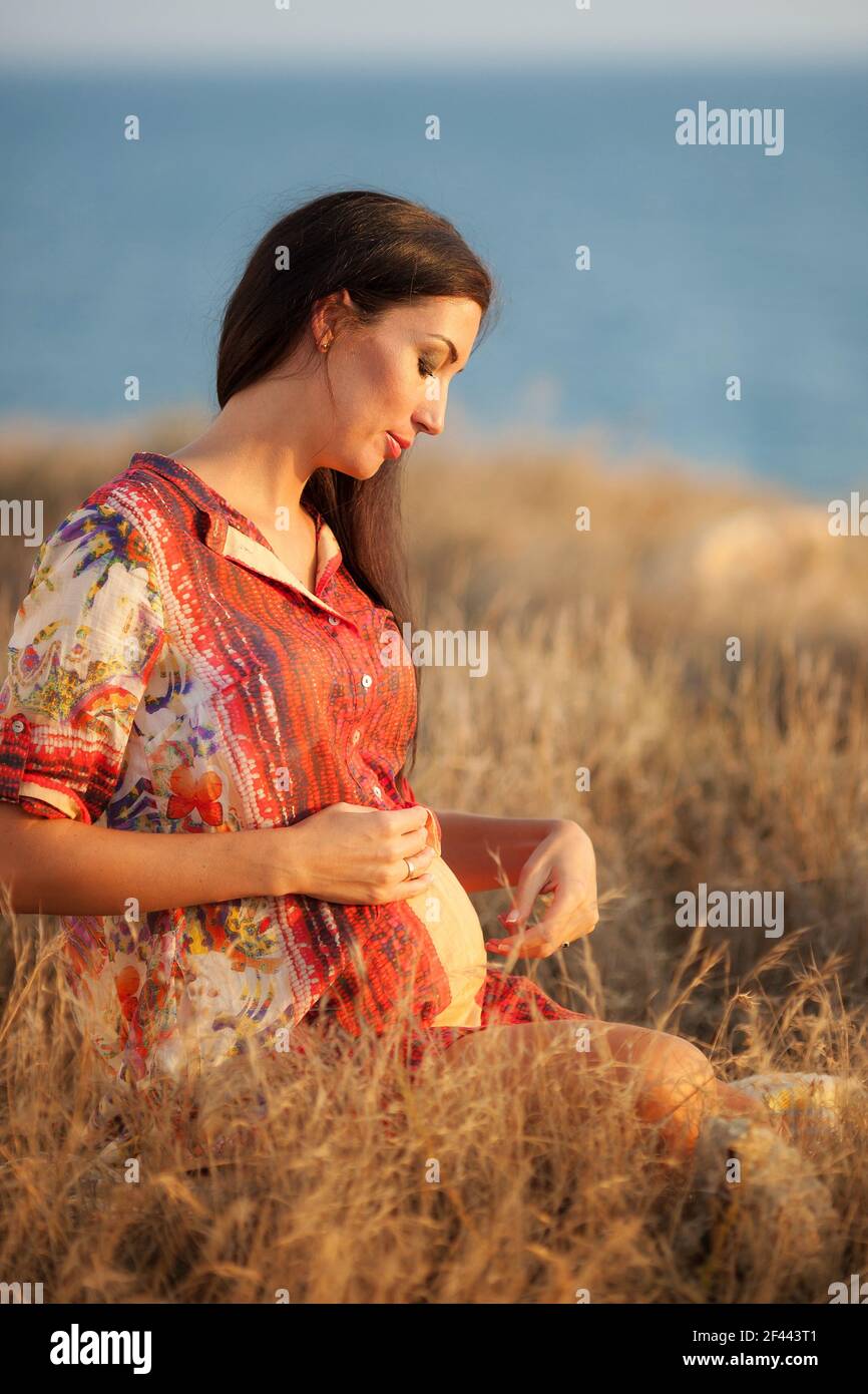Schöne Schwangeren entspannen draußen in der Nähe des Meeres. Glückliche Schwangere mit langen Haaren genießen das Leben in der Natur. Stockfoto