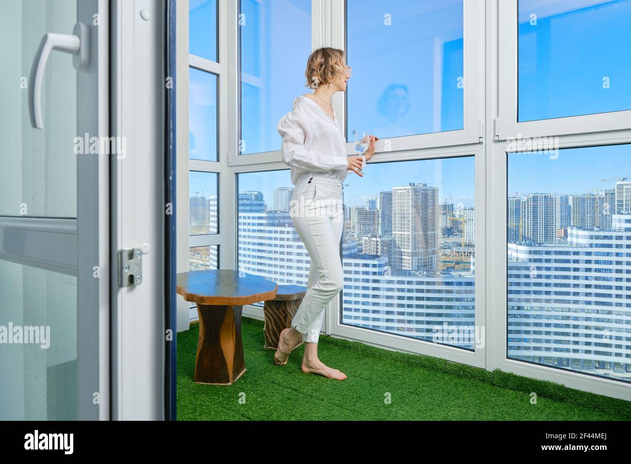 Barfußfrau steht auf dem Balkon ihrer neuen Wohnung Und bewundert die Aussicht auf die Stadt aus dem Fenster Stockfoto