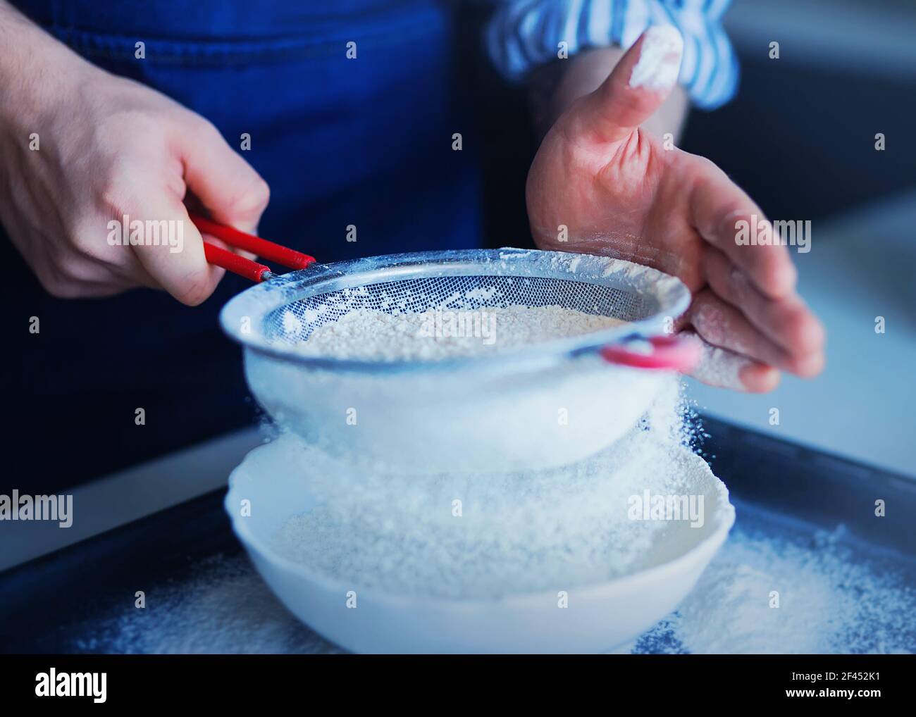 Ein Mann in einer blauen Schürze siebt Mehl durch ein Sieb in eine weiße Schüssel, in der er die Zutaten für den Teig mit dem Mehl mischen wird. Home Co Stockfoto