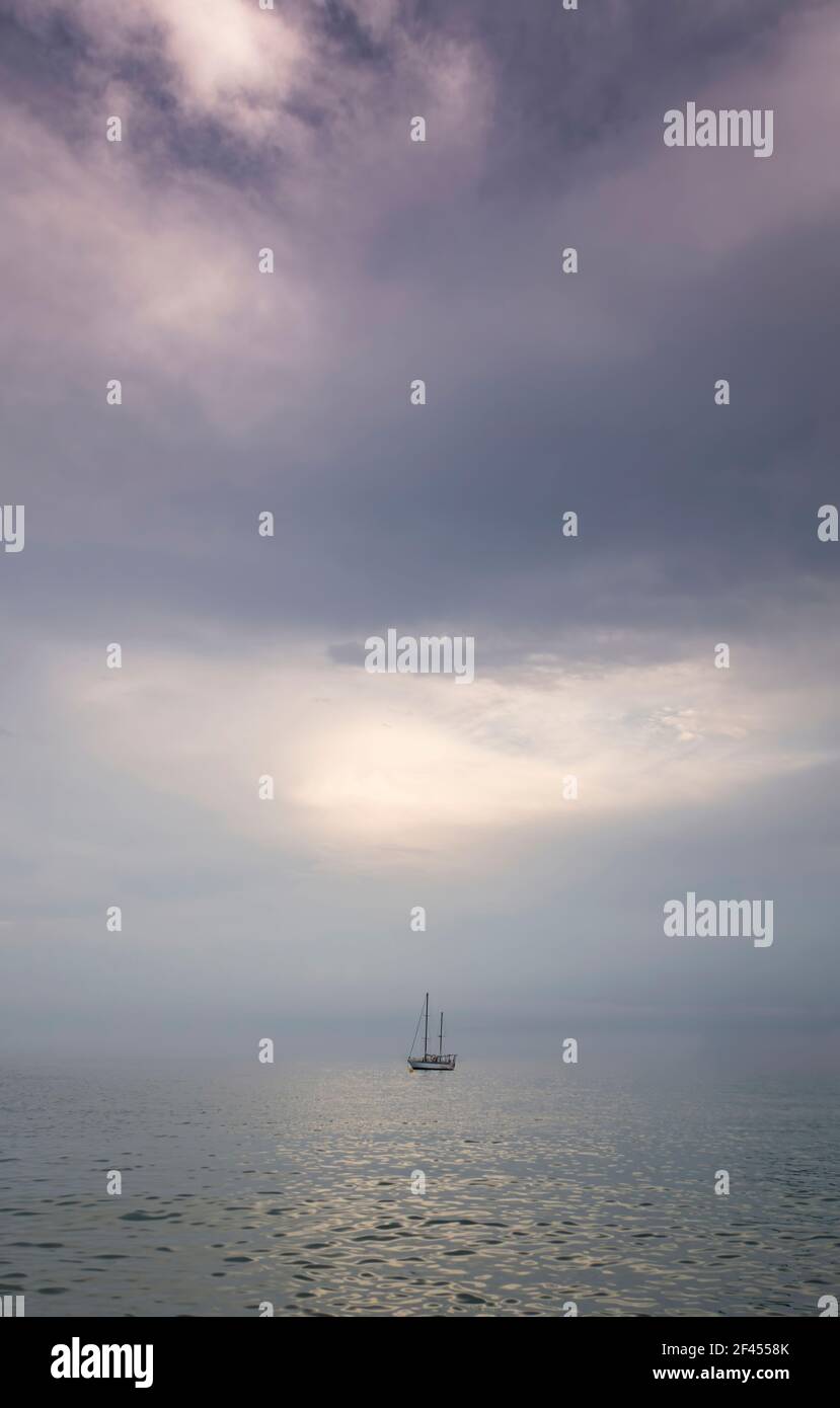 Ein kleines Segelboot in der Mitte eines Meeres in völliger Ruhe in einem Sonnenuntergang mit einem bewölkten Himmel in mauve Töne, Gefühl der Ruhe, Isolation, Minimalismus und verankert Stockfoto