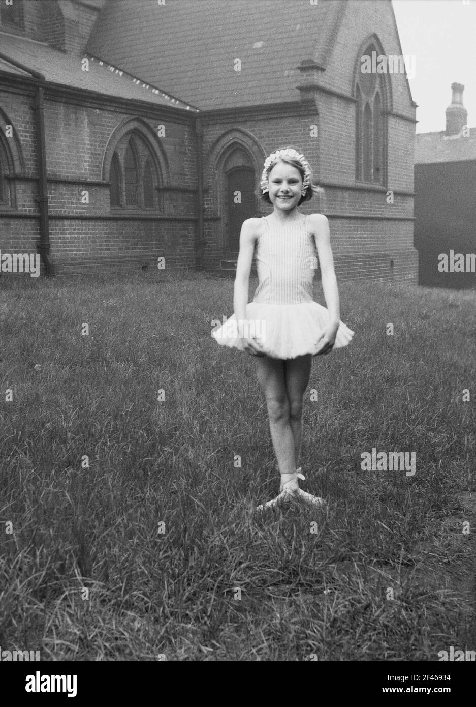 1956, historisch, ein junges Mädchen, das draußen auf dem Gras auf dem Gelände einer Kirche in einem kurzen Ballettkleid steht, bevor es an der Parade zum 1. Mai teilnimmt, einem beliebten Feiertag, der den Beginn des Sommers markiert. May First, allgemein bekannt als May Day, ist eine alte Tradition, die auf die Feier der römischen Blumengöttin Flora zurückgeht. Stockfoto