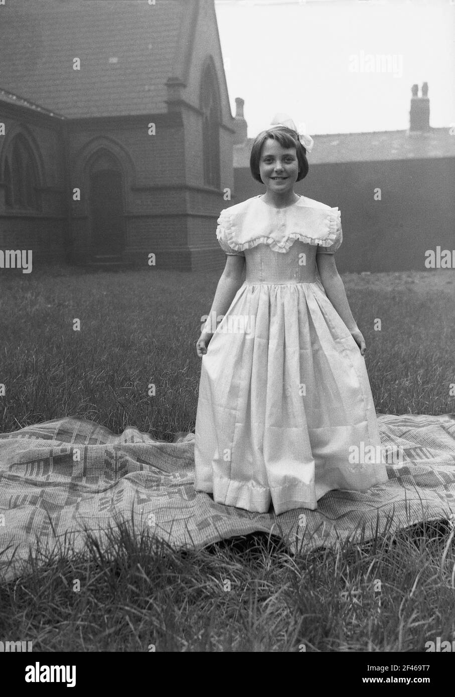 1956, ein junges Mädchen im Teenageralter, das draußen auf dem Gelände einer Kirche mit einem langen, aufwendigen Spitzenkleid auf einem Teppich steht, bevor es an der Parade zum 1. Mai teilnimmt, einem beliebten Feiertag, der den Beginn des Sommers markiert. May First, allgemein bekannt als May Day, ist eine alte Tradition, die auf die Feier der römischen Blumengöttin Flora zurückgeht. Stockfoto