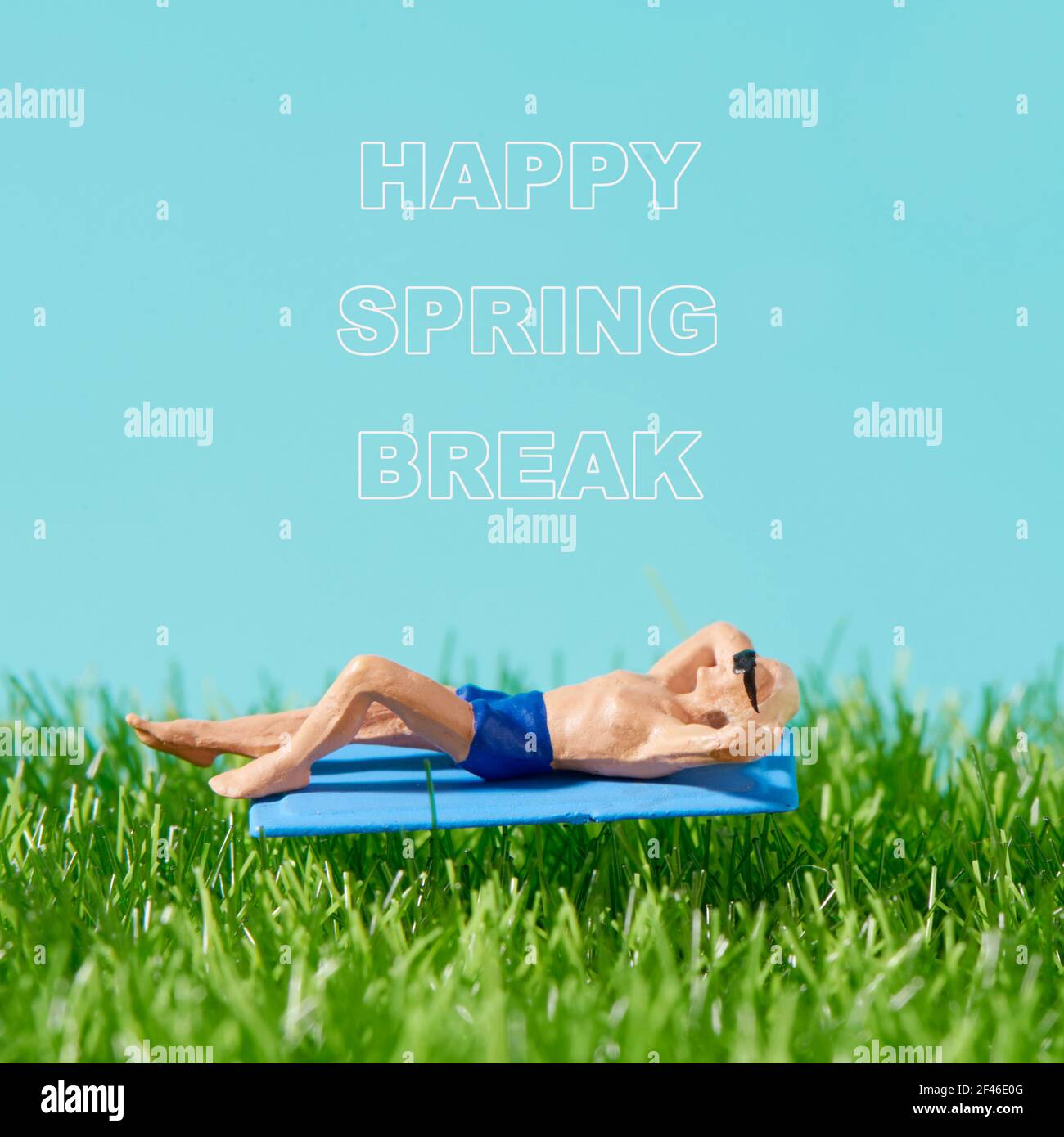Nahaufnahme eines Miniaturmannes, der Badeanzug und Sonnenbrille trägt, sich auf einem blauen Handtuch auf dem Gras legt, und der Text Happy Spring Break auf dem blauen Rücken Stockfoto