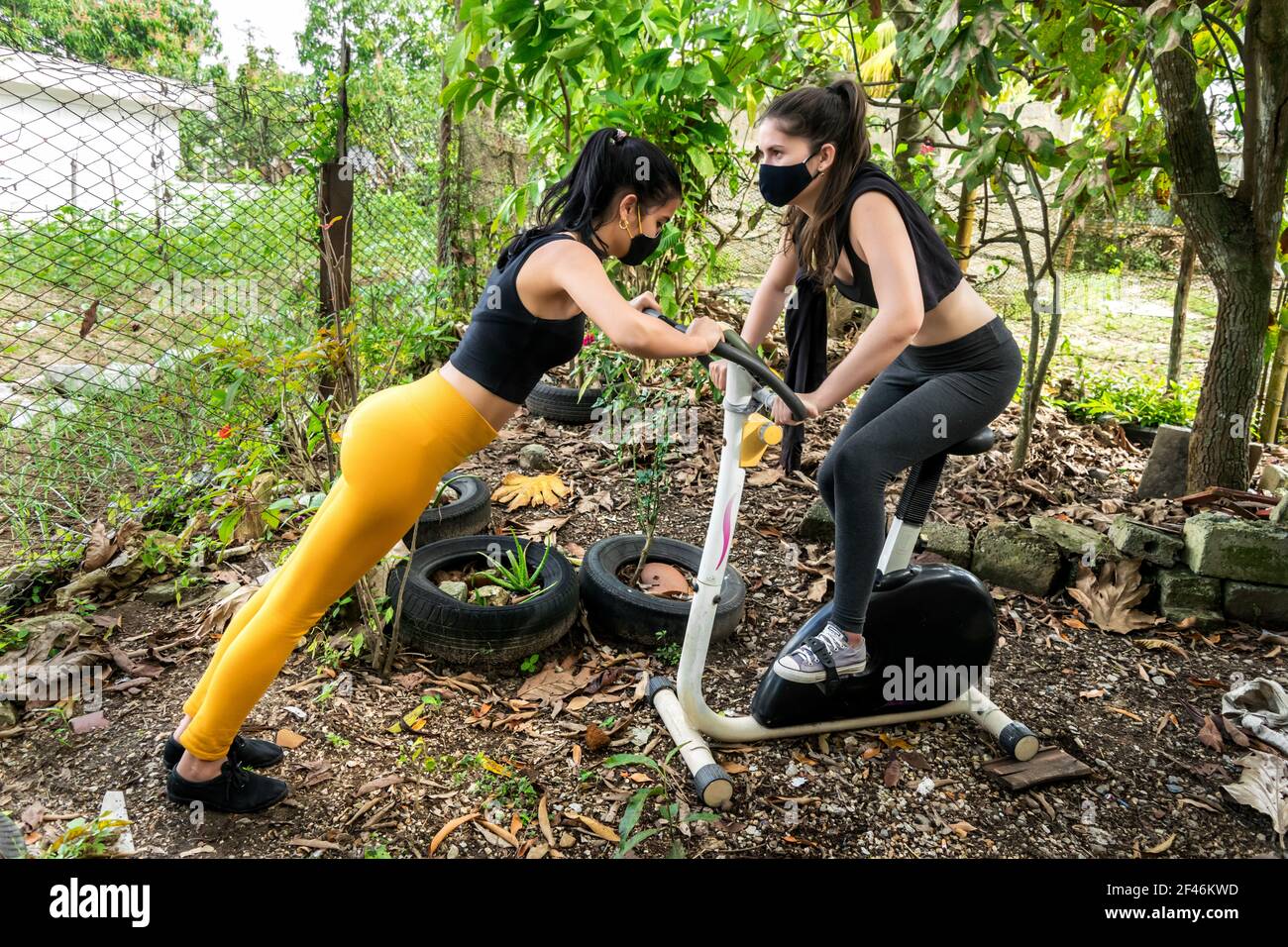 Zwei junge kubanische Frauen arbeiten zusammen draußen in einem verlassenen Hinterhof, sie sind mit einem statischen Fahrrad Stockfoto