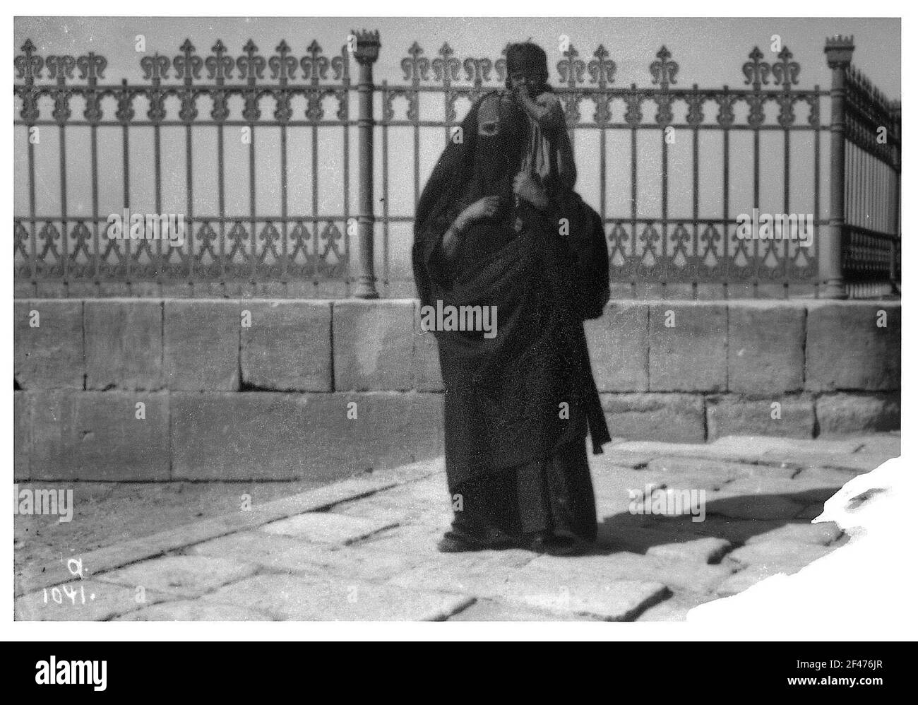 Indien (?). Verschleierte Frau mit Kleinkind auf der Schulter vor einem eisernen Gitterzaun Stockfoto