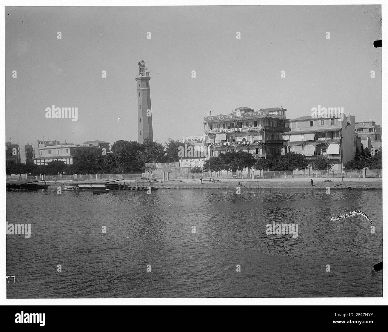 Port Said, Ägypten. Blick von einem Hochsee-Passagierdampfer von Hapag auf Uferstreifen des Suez-Kanals mit Bootsanlegestelle, Leuchtturm und Werbung für englische Produkte Stockfoto