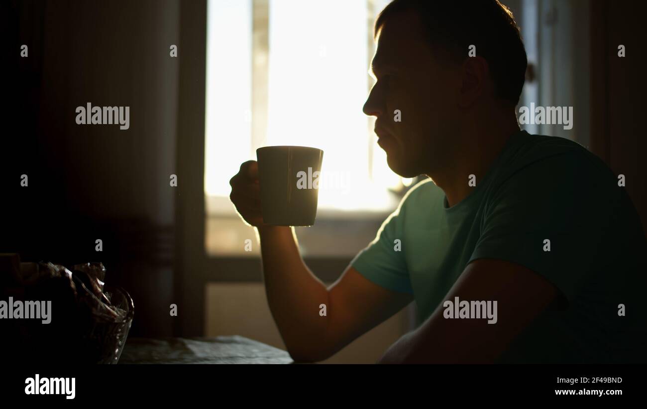 Silhouette eines Mannes in den Sonnenstrahlen der Morgendämmerung trinkt heißen Kaffee, während zu Hause sitzen, Kamerabewegung Stockfoto