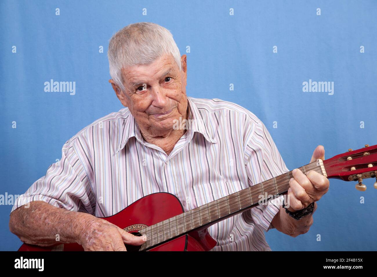 Aktiver glücklicher älterer Mann mit grauem Haar und einer sonnengeschädigten Haut, der auf einer roten Gitarre vor blauem Hintergrund Musik spielt. Stockfoto Aktiver glücklicher älterer Mann mit grauem Haar und einer sonnengeschädigten Haut, der auf einer roten Gitarre vor blauem Hintergrund Musik spielt. Stockfoto