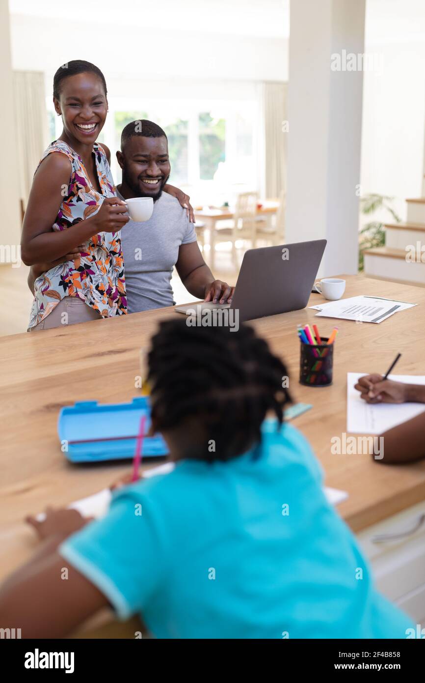Glückliche afroamerikanische Eltern mit Laptop umarmt halten Kaffee mit Kinder arbeiten am Tisch Stockfoto
