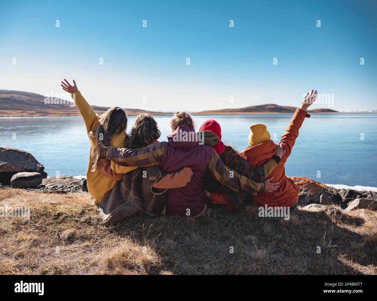 Eine Gruppe von glücklichen jungen Freunden sitzen in der Nähe des Bergsees, umarmen und Spaß haben Stockfoto