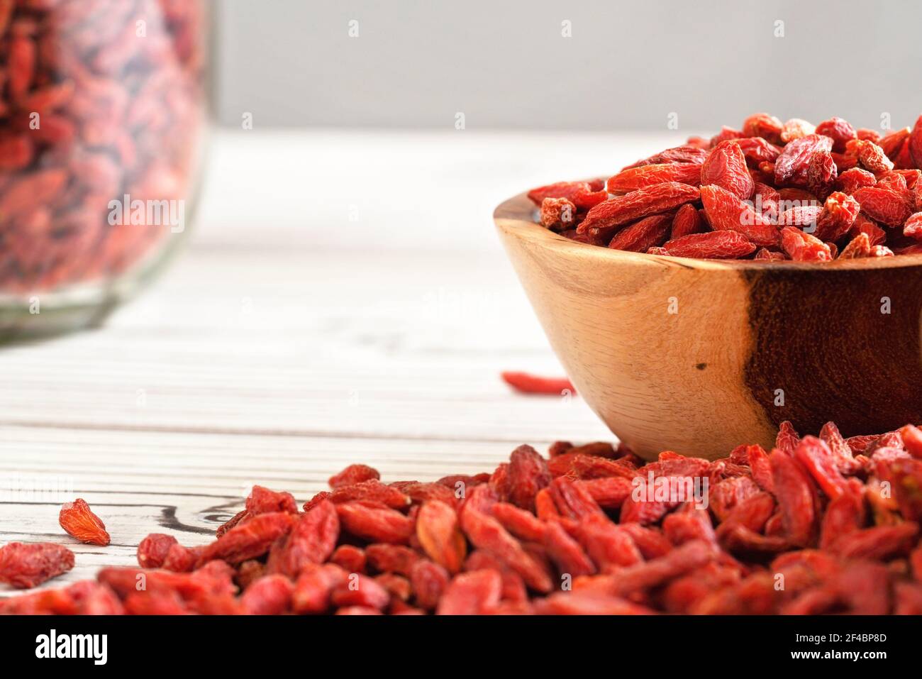Getrocknete Goji Beeren in Holzschale, verstreut über weißen Brettern Tisch unter, verschwommen großen Glas voller Früchte im Hintergrund Stockfoto
